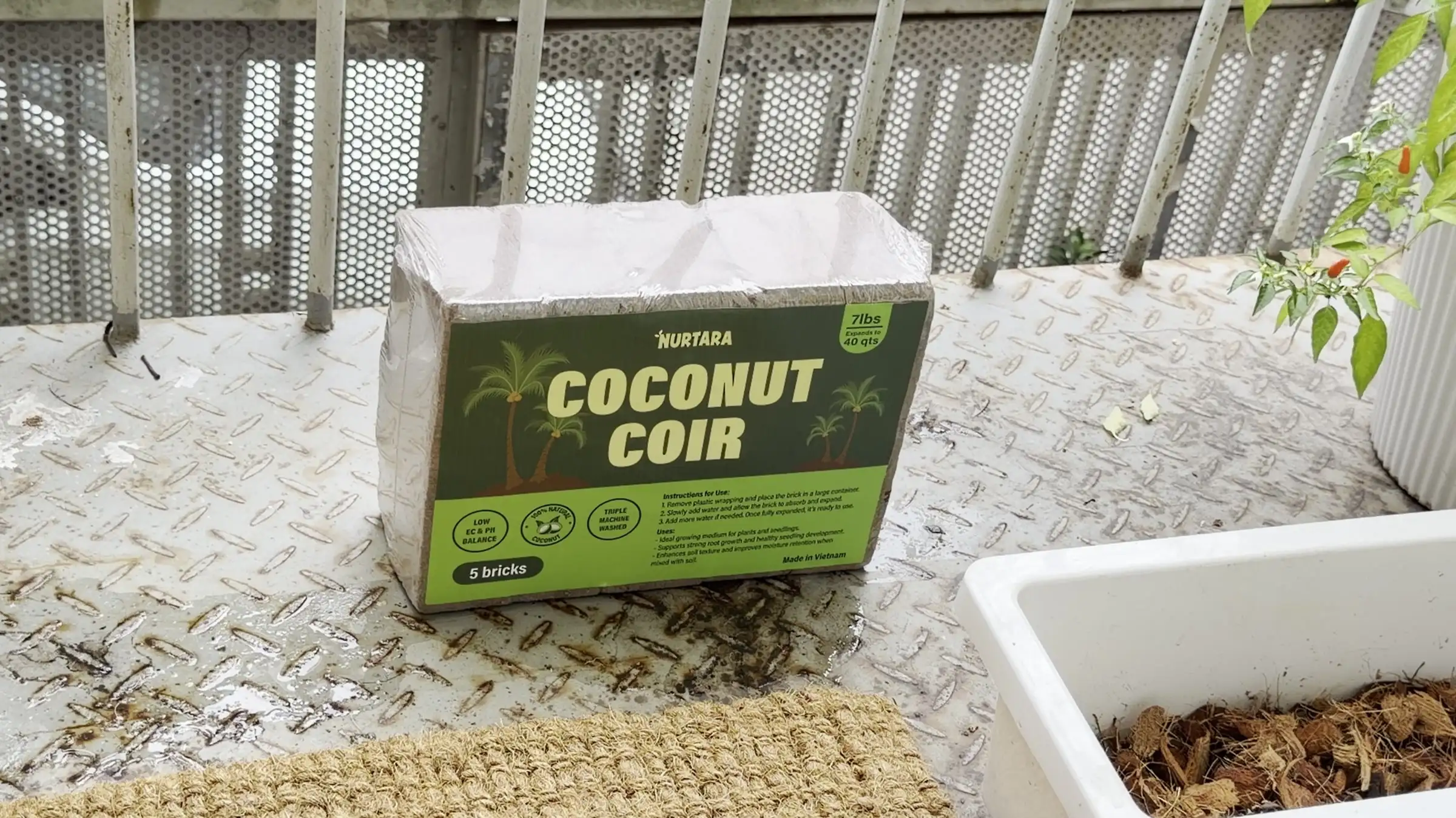 A NURTARA Coconut Coir pack and an unwrapped compressed coir brick next to a white planter on a balcony